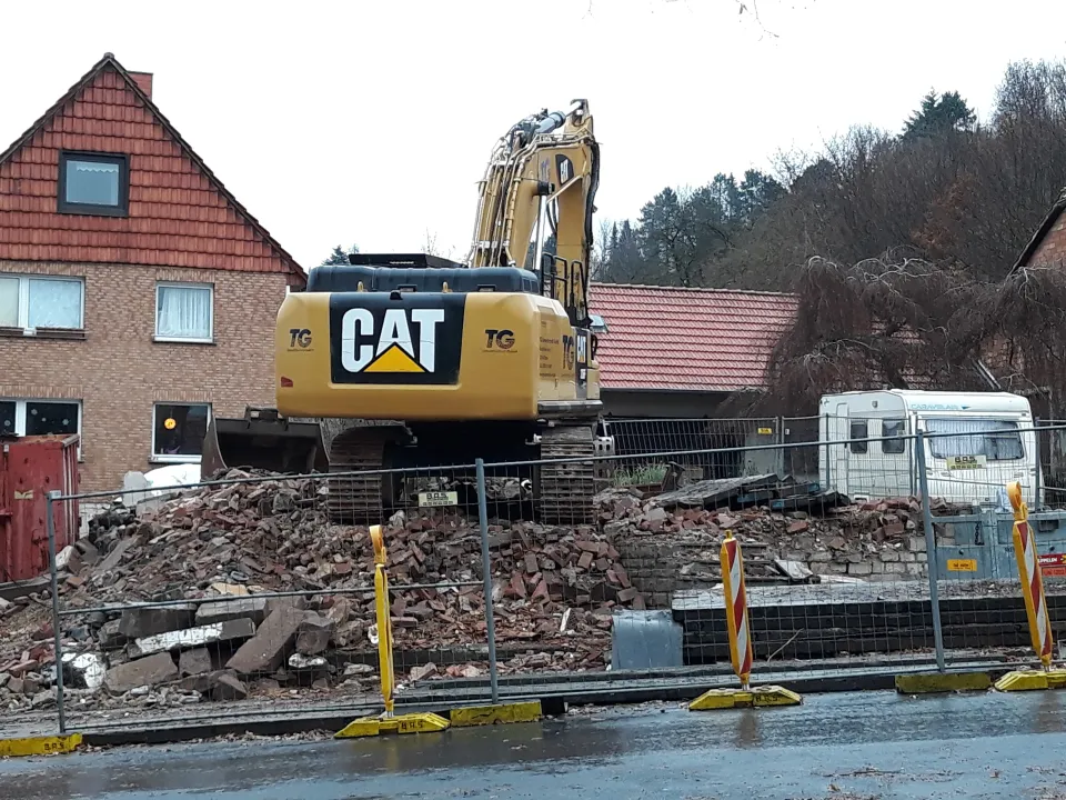 Abrissbagger vor einem Schutthaufen hinter dem Bauzaun an der Straße, links ein Backsteinhaus, rechts ein Wohnwagen; vom Haus ist nur noch der Schutt übrig.