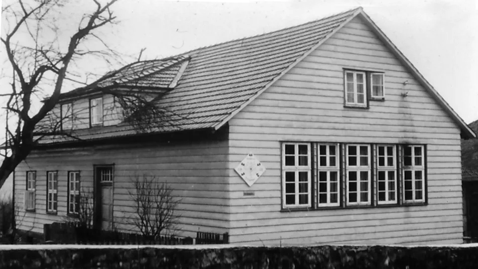 Historisches Schwarzweißfoto der alten Dorfschule: zweigeschossiges Haus mit Satteldach und Gaube, mit waagerechten Brettern verschalt, an der Giebelseite fünf Fenster in einer Reihe und eine rautenförmige Tafel; davor eine Bruchsteinmauer.