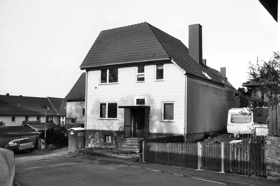 Schwarzweißfoto eines freistehenden Wohnhauses mit Walmdach, hellem Brettbehang und Steinsockel, an der Haustür steht die Hausnummer 27; davor ein Bauzaun, rechts ein Wohnwagen.