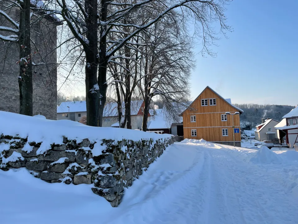 Verschneiter Weg entlang einer Bruchsteinmauer mit Schneehaube, links der Kirchturm und kahle Bäume, rechts im Hintergrund ein Haus mit gelber Holzverschalung und weitere Wohnhäuser unter blauem Himmel.