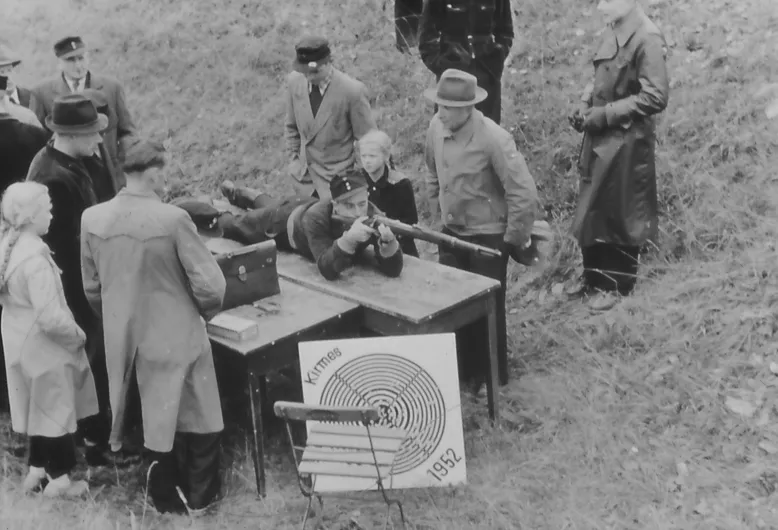 Schwarzweißfoto: Ein Schütze liegt auf einem Tisch und zielt mit einem Gewehr, daneben stehen Zuschauer in Mänteln und Hüten; vorn lehnt eine Scheibe mit der Aufschrift „Kirmes 1952“.