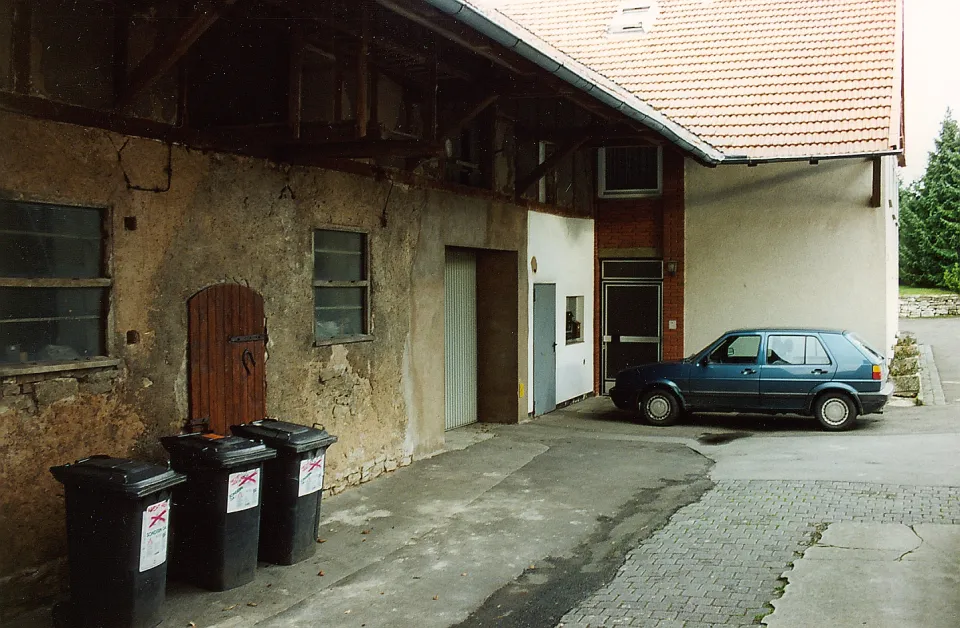 Hofseite des Gasthauses mit Nebengebäuden vor dem Umbau: links eine alte Lehmwand mit Holztür und drei Mülltonnen, rechts ein hell verputzter Bau mit rotem Ziegeldach, davor ein geparkter Pkw auf Pflaster.