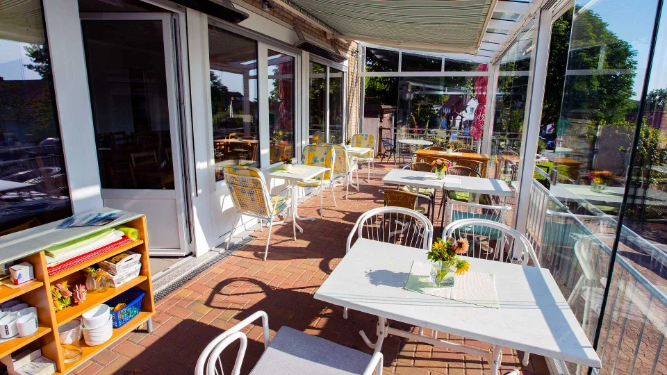 Gedeckte Tische mit Blumenschmuck auf einer gepflasterten Terrasse mit Glaswänden und Markise, Stühle mit gelben Kissen, links ein Regal mit Geschirr.