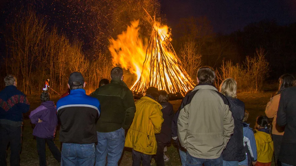Menschen, darunter Kinder, von hinten vor einem brennenden kegelförmigen Holzstoß bei Nacht, Funken steigen auf.