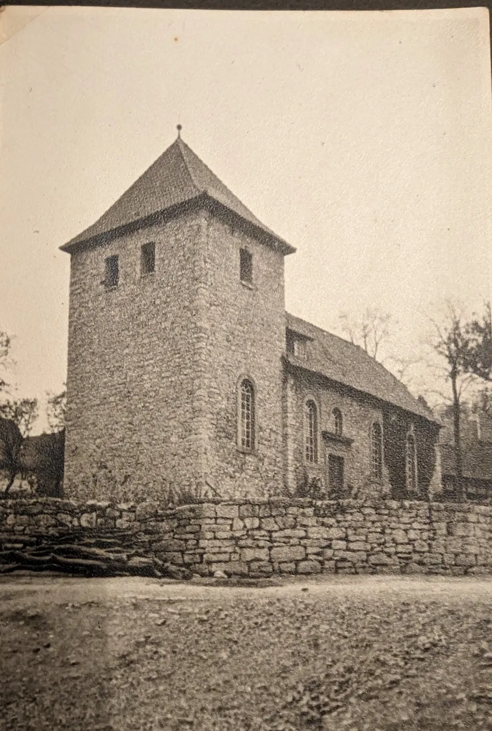 Schwarzweißfoto der St.-Martins-Kirche: links der wuchtige Westturm aus Bruchstein mit Pyramidendach und schmalen Schlitzfenstern, rechts das niedrigere Schiff mit Rundbogenfenstern, davor eine Bruchsteinmauer.